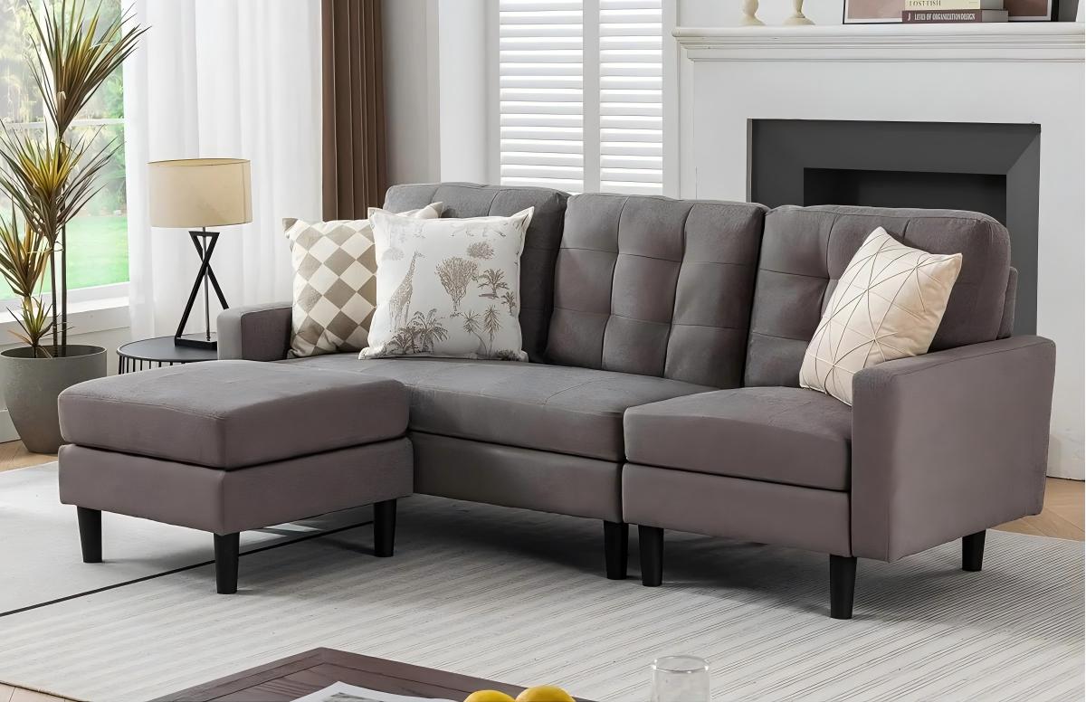 Modern gray L-shaped sectional sofa with tufted cushions and patterned throw pillows in a bright living room with large windows and neutral decor.