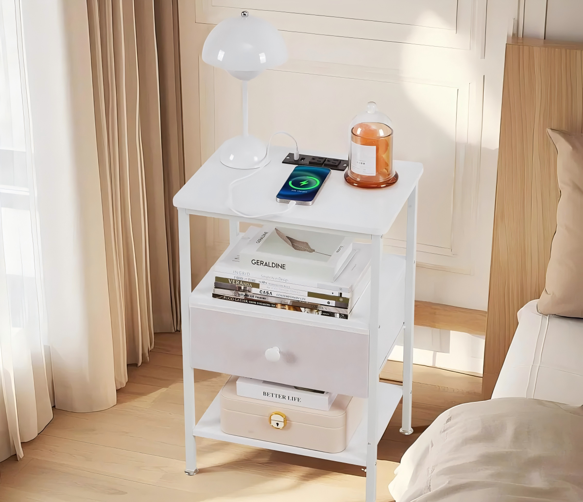 A compact white nightstand with built-in charging ports, a drawer, and open shelves, neatly organized with books and accessories beside a bed in a softly lit room.