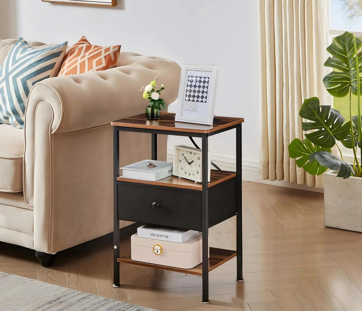 Industrial-style side table with wood-look shelves, a single drawer, and black metal frame, placed beside a beige sofa and styled with a photo frame, small plant, clock, and storage box in a bright living room.