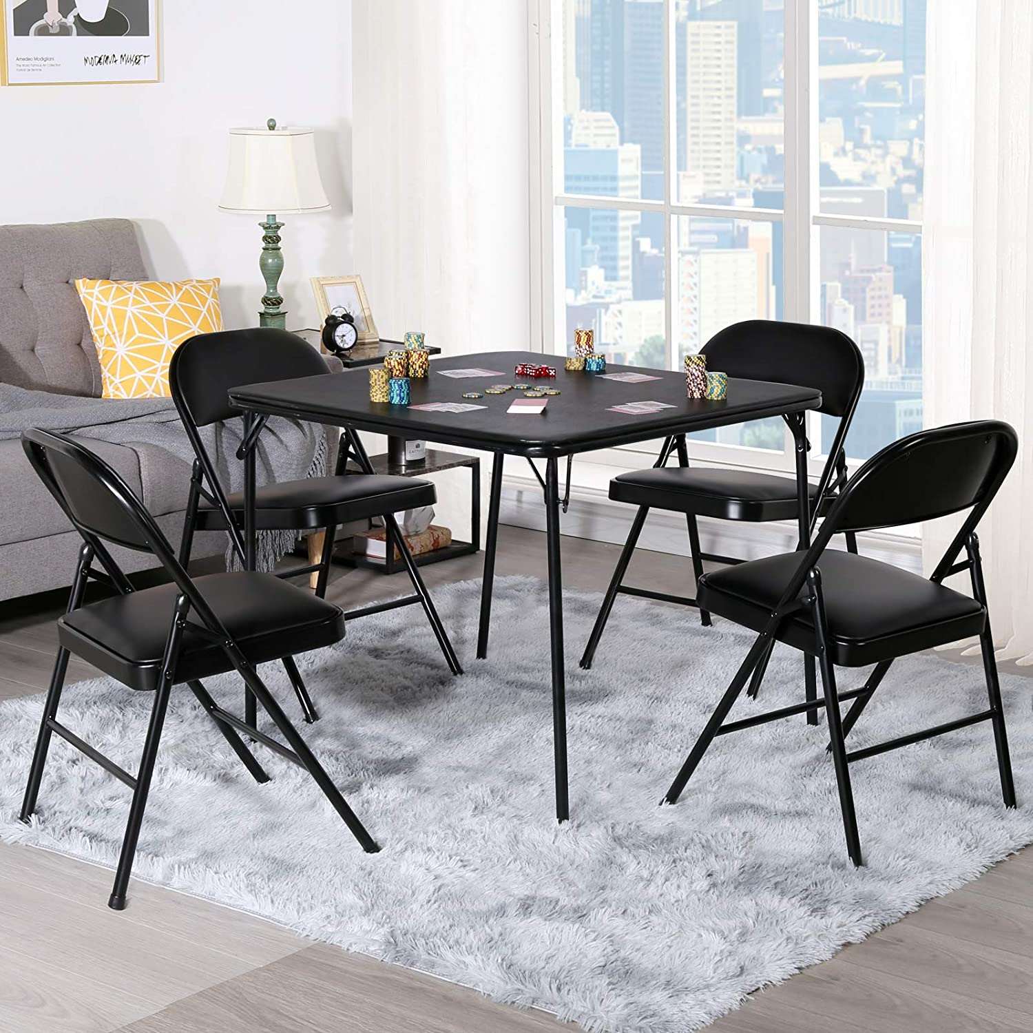 Black square folding card table with four matching padded folding chairs, set up for poker with chips and cards in a bright living room.
